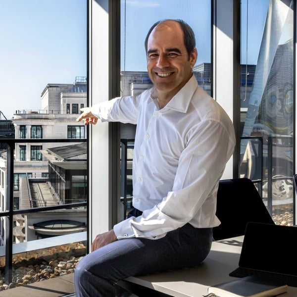 António Simões sitting on a desk in L&G's office at 1 Coleman Street, with a large office window behind him, pointing towards 10 Coleman Street.