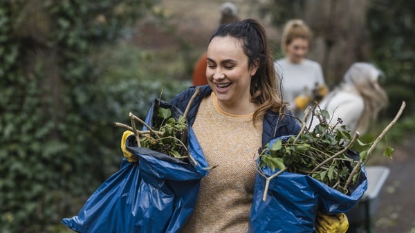 A person in an outdoor park is holding two blue plastic bags filled with plant debris. In the background, another individual is pushing a wheelbarrow.