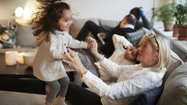 A young girl plays with her grandmother on the sofa, surrounded by her family at home.