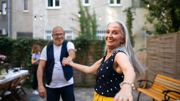 A senior woman dancing with her husband outdoors in a paved garden.