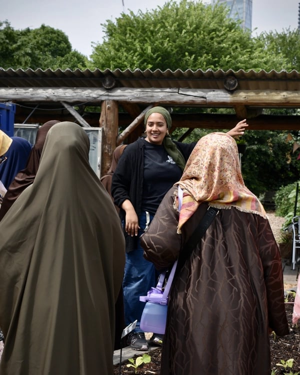 A group of women in hijabs listen to a speaker outdoors, with trees and a wooden structure in the background.