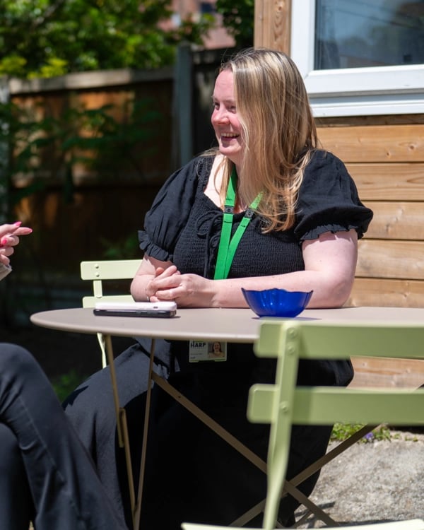 Two people with blurred faces sit at a round outdoor table talking. Both wear green lanyards; a blue bowl and smartphone are on the table. Greenery and a wooden fence are in the background.