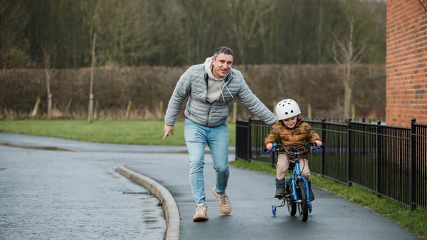 A father rests a reassuring hand on his young son's back as he learns to ride his bike on the footpath outside of his home in the North East of England. The young boy is smiling as he controls the bike on the footpath.
