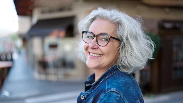 Grace, a woman with short grey hair and glasses, stands outdoors, smiling as she looks at the camera.