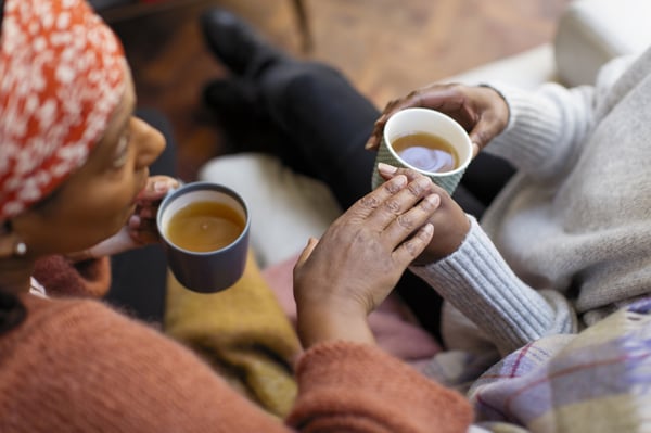 Two people sitting together indoors, holding cups of tea, with one person resting a hand on the other's arm in a supportive gesture.