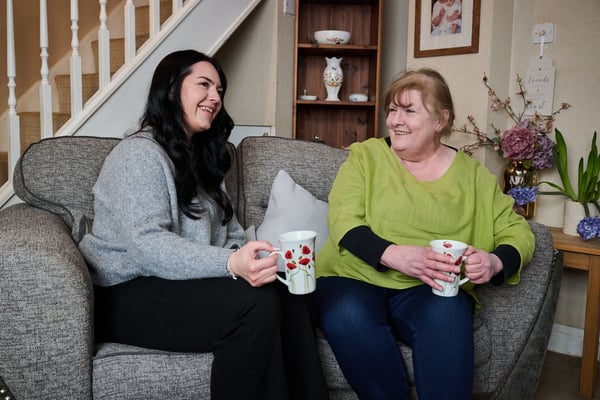 Two people sitting on a sofa in a living room, holding mugs and talking.
