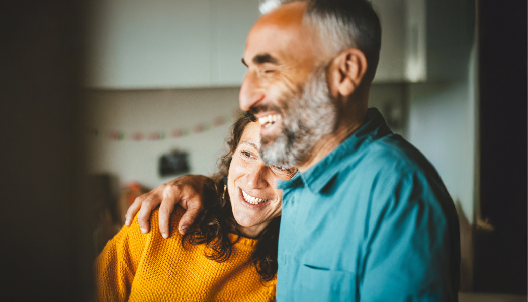 A person in a teal shirt with an arm around another person wearing a yellow sweater inside a softly lit kitchen.