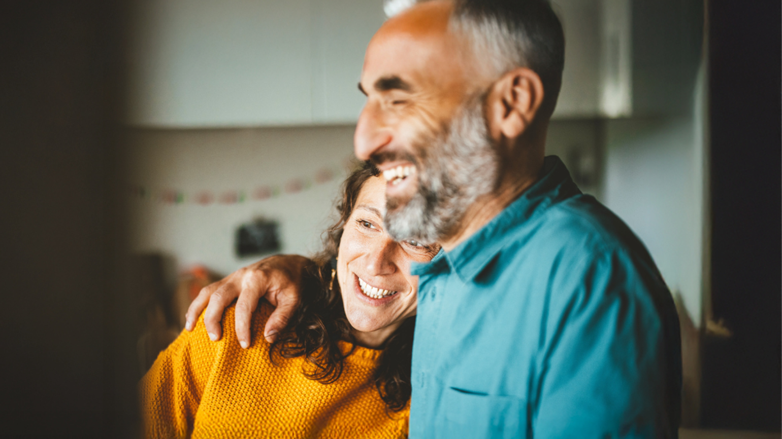 A person in a teal shirt with an arm around another person wearing a yellow sweater inside a softly lit kitchen.