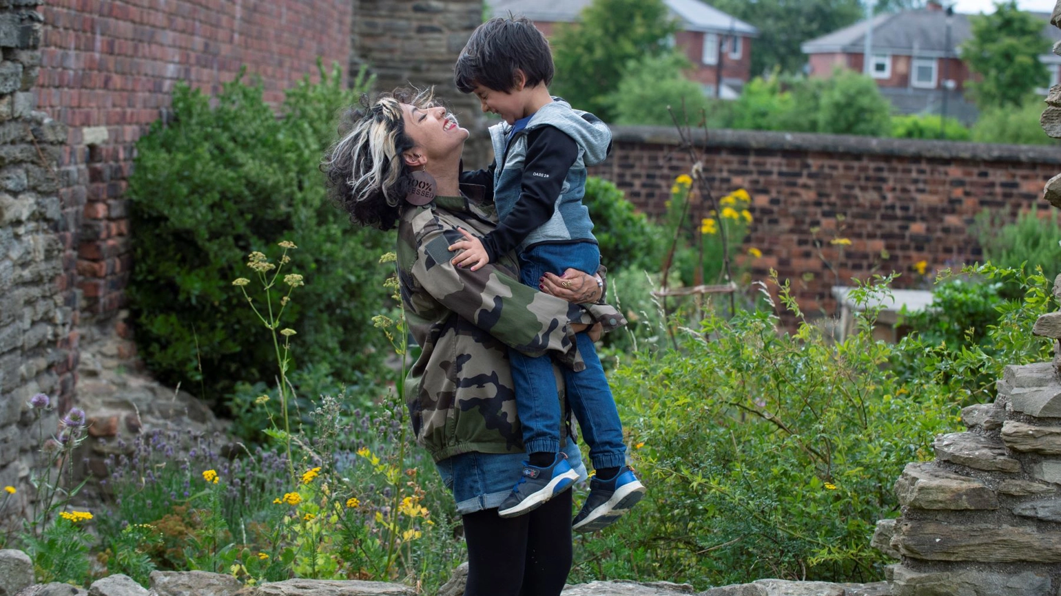A beaming women carries a child, the pair are in a garden that has wildflowers and rockery A beaming women carries a child, the pair are in a garden that has wildflowers and rockery