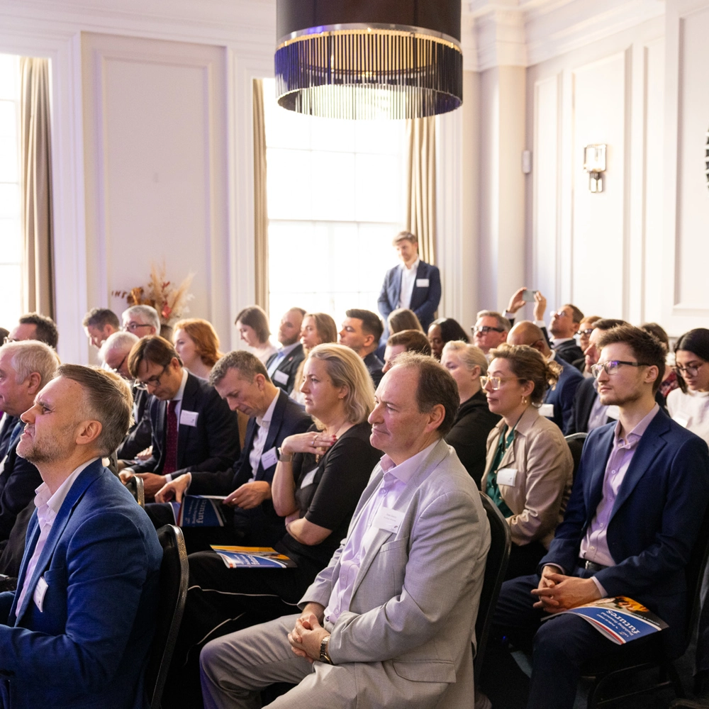 Attendees seated closely together reading brochures and event materials during a session. Attendees seated closely together reading brochures and event materials during a session.