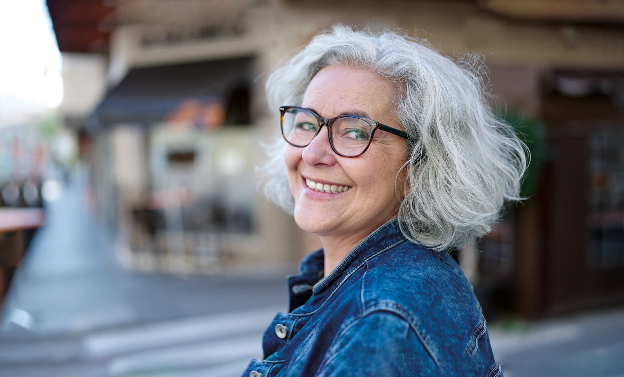 Grace, a woman with short grey hair and glasses, stands outdoors, smiling as she looks at the camera.