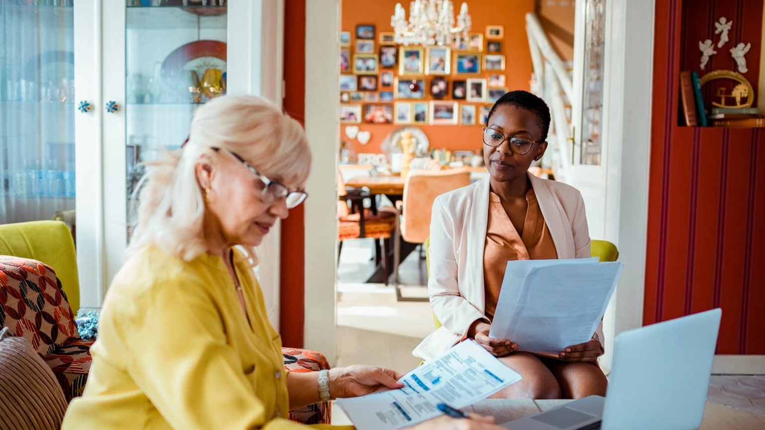 Two women working on their financial accounts, with one seated on a sofa and the other on a chair, both focused on reviewing paperwork. Two women working on their financial accounts, with one seated on a sofa and the other on a chair, both focused on reviewing paperwork.