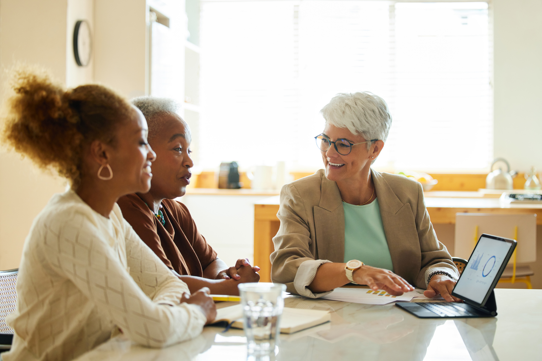 Three people seated at a table in a modern, in a well-lit room, engaged in discussion. A tablet is placed on the table in front of one person, along with documents and glasses of water. The background features a sofa and framed artwork of the walls.