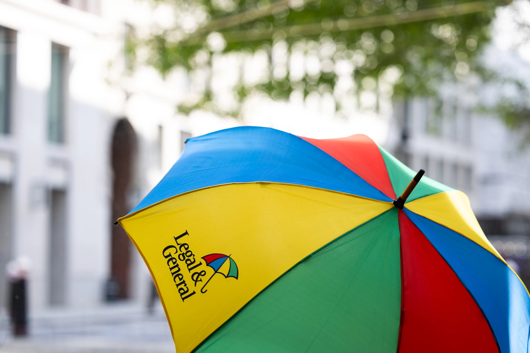 A colorful umbrella with panels in blue, green, yellow, and red. Our Legal & General logo is displayed on one of the yellow panels. The background is blurred, featuring buildings and trees.