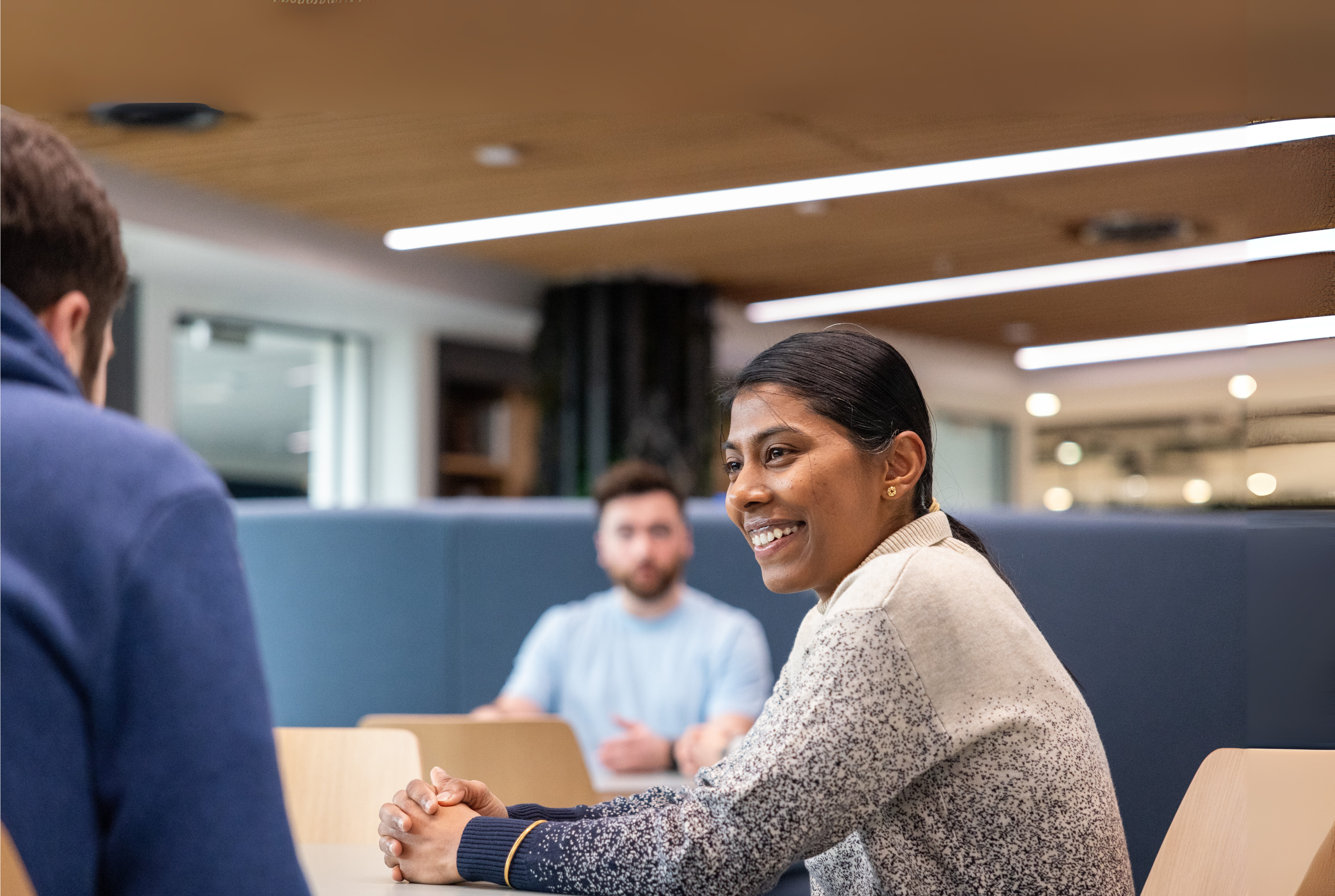 Two colleagues are seated around a table in a well-lit room with wooden ceiling panels. One wears a white and blue speckled jumper, another a navy blue hoodie.,