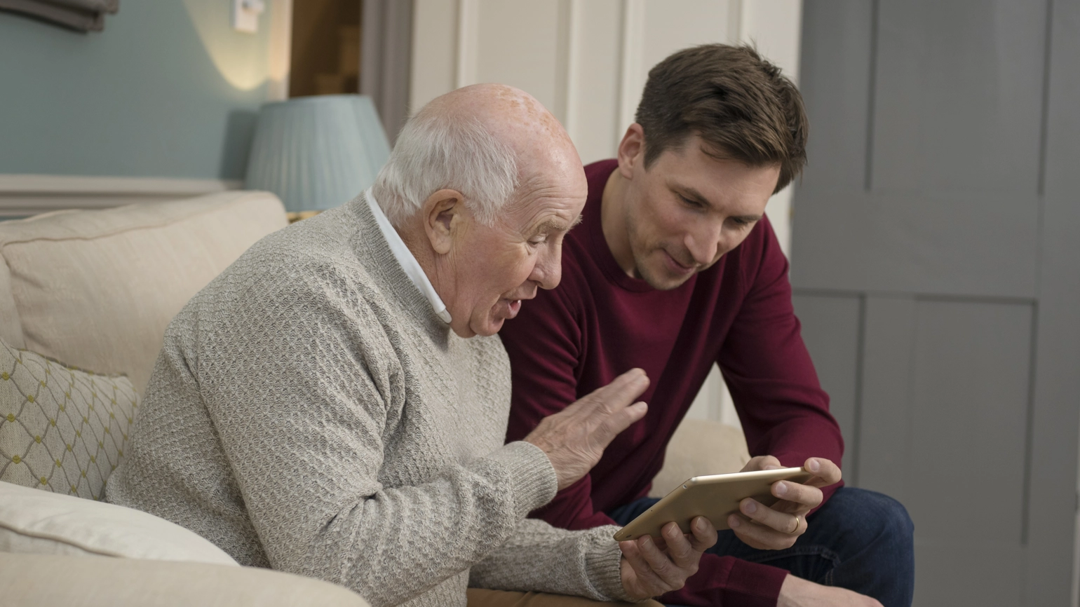 Middle aged male and elderly male chatting with their touch screen in living room Middle aged male and elderly male chatting with their touch screen in living room