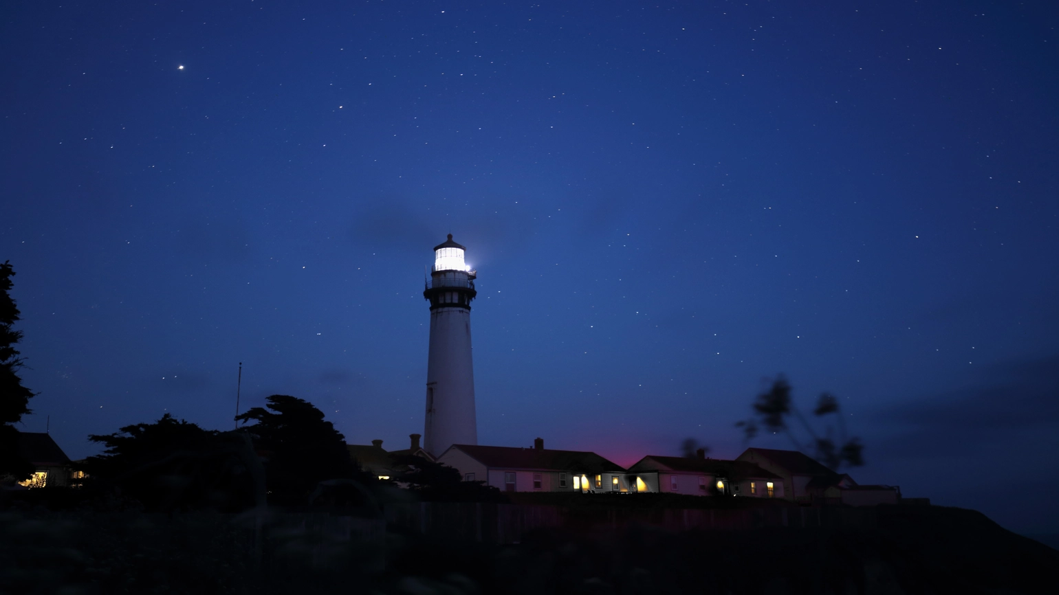 Pigeon Point Lighthouse at night, its beacon shining brightly over nearby buildings with lit windows. Pigeon Point Lighthouse at night, its beacon shining brightly over nearby buildings with lit windows.