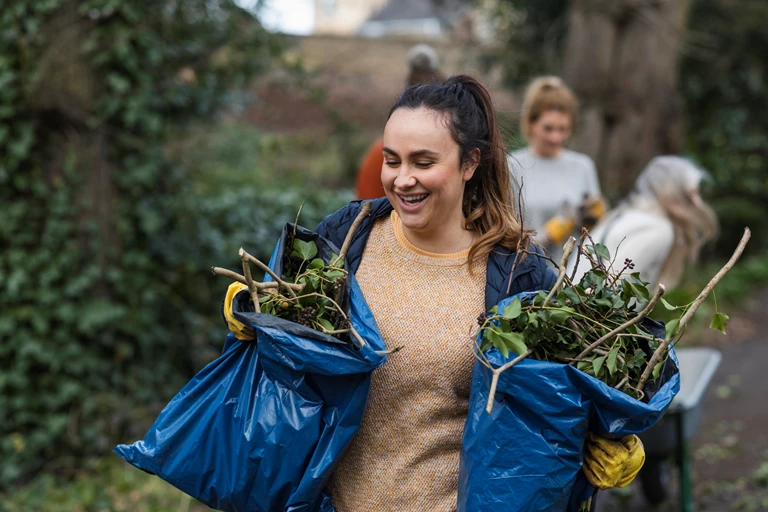 A person in an outdoor park is holding two blue plastic bags filled with plant debris. In the background, another individual is pushing a wheelbarrow. 