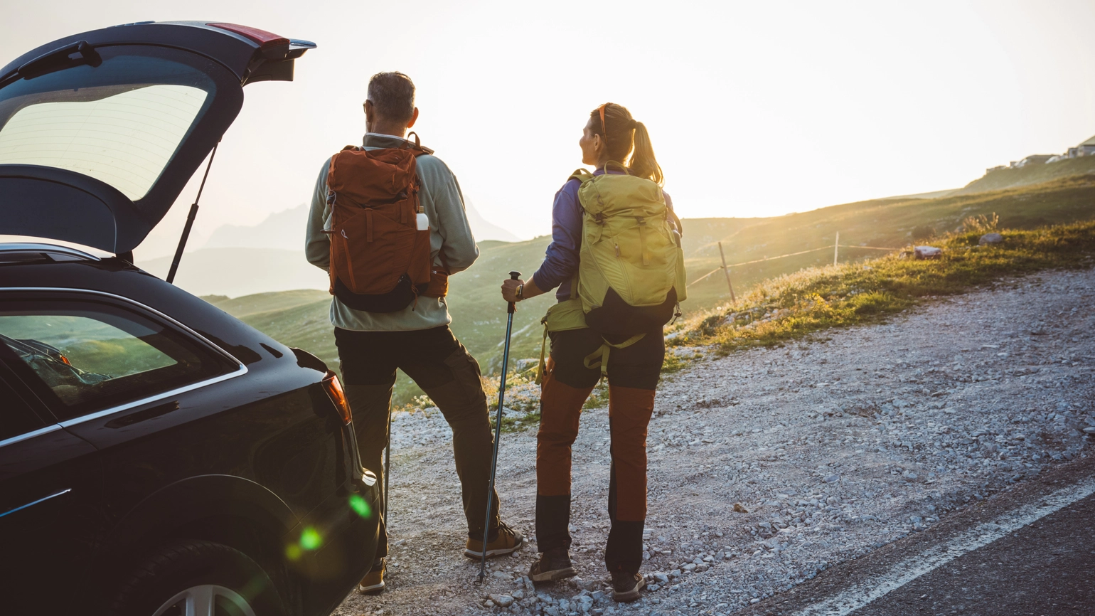 Two hikers standing beside an open car trunk on a gravel roadside, wearing backpacks and outdoor gear, looking toward sunlit grassy slopes and mountain peaks in the distance. Two hikers standing beside an open car trunk on a gravel roadside, wearing backpacks and outdoor gear, looking toward sunlit grassy slopes and mountain peaks in the distance.