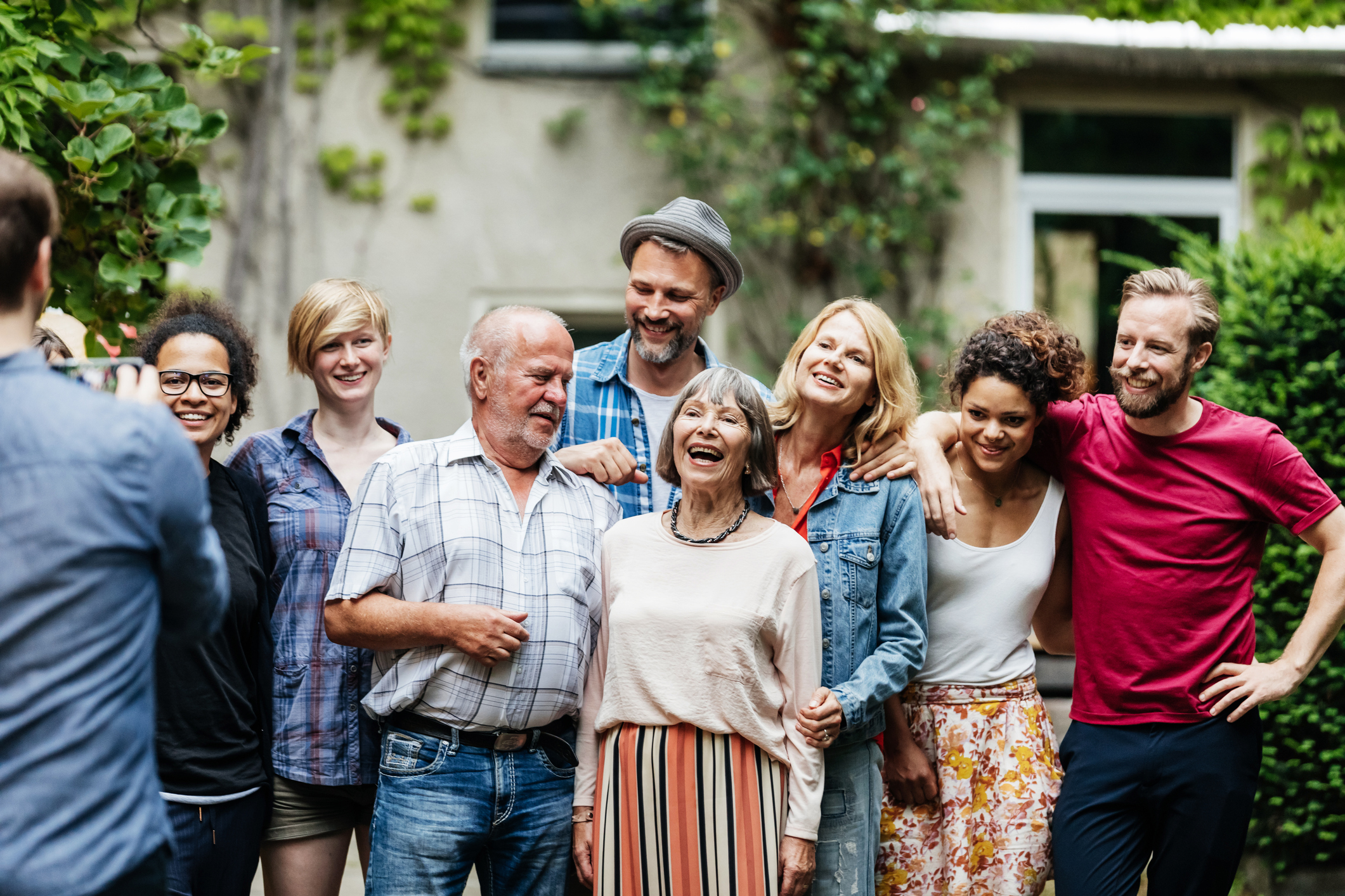 A group of people gathered outside in a leafy back garden, standing close together for a photo. They’re dressed casually, with some resting their arms on each other’s shoulders. The background shows part of a house with windows.