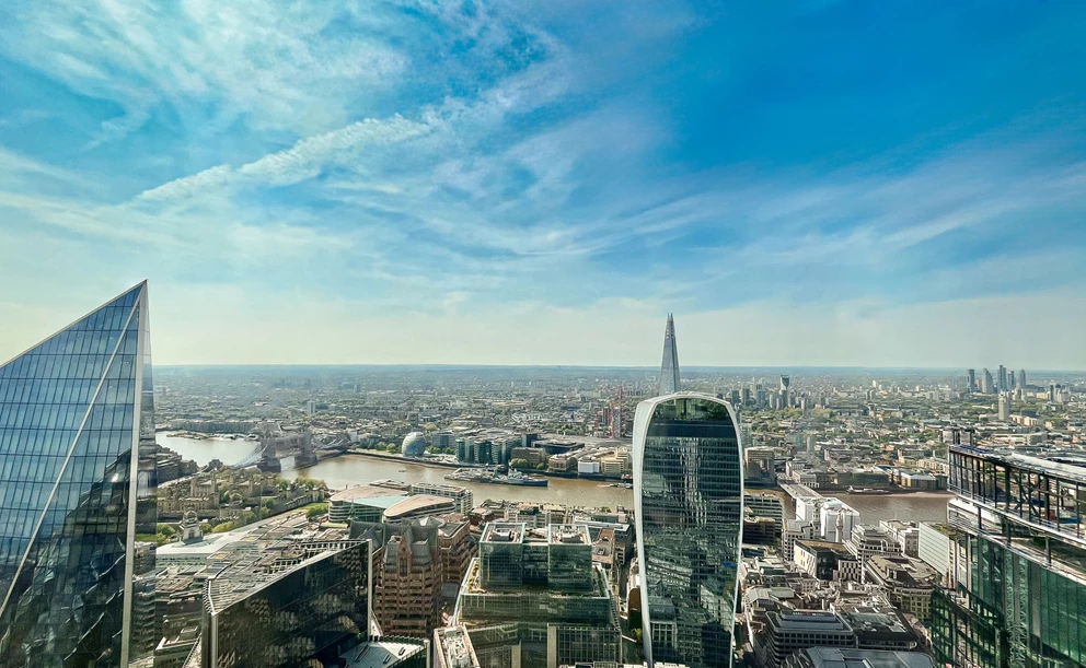 A high-angle view of London under clear blue skies. There is a mix of modern skyscrapers with reflective glass facades, like the Fenchurch Building (also known as The Walkie-Talkie). The River Thames winds through the photograph.