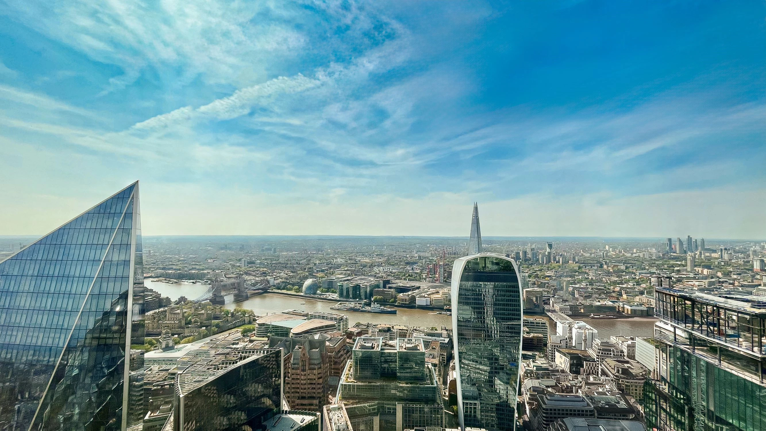 A high-angle view of London under clear blue skies. There is a mix of modern skyscrapers with reflective glass facades, like the Fenchurch Building (also known as The Walkie-Talkie). The River Thames winds through the photograph.