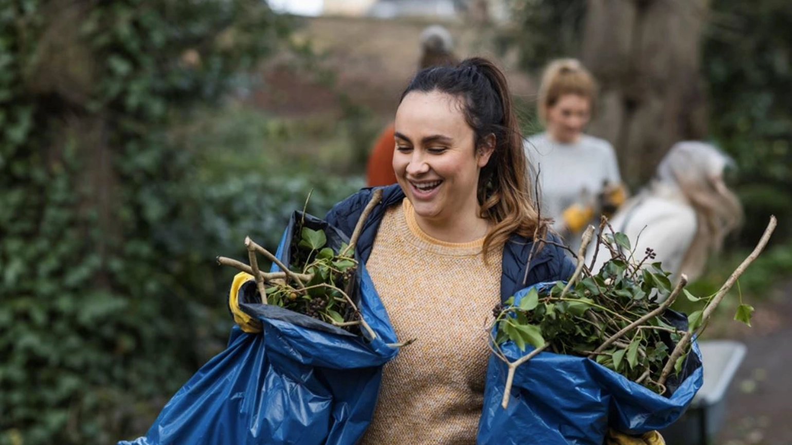 A volunteer tidying a garden, carrying two bags of green waste. A volunteer tidying a garden, carrying two bags of green waste.