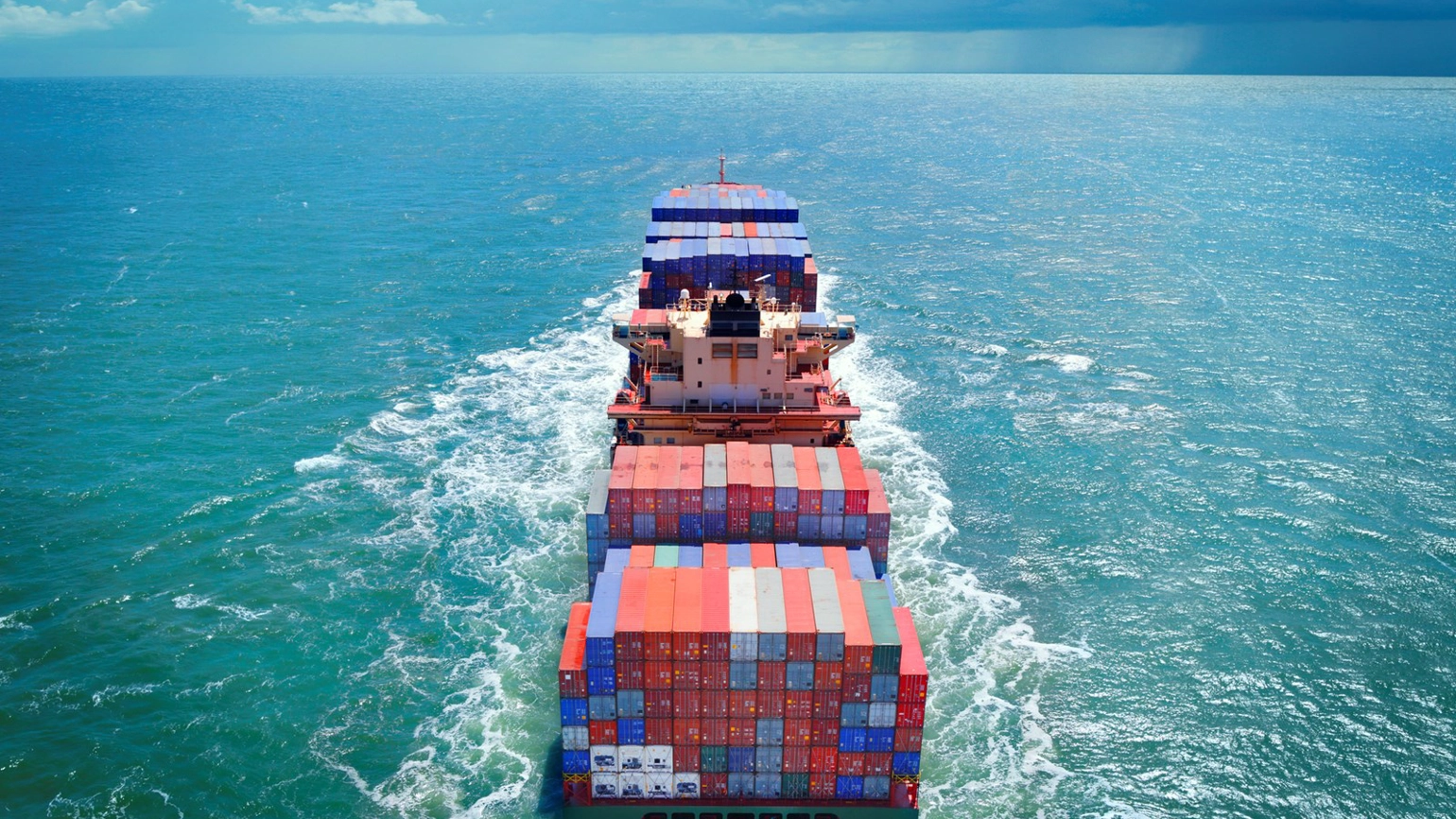 Aerial view of a large freight ship carrying red, blue, and white cargo containers, sailing through the open sea. The ship creates a wave as it moves forward, with water stretching to the horizon under a partly cloudy sky. Aerial view of a large freight ship carrying red, blue, and white cargo containers, sailing through the open sea. The ship creates a wave as it moves forward, with water stretching to the horizon under a partly cloudy sky.