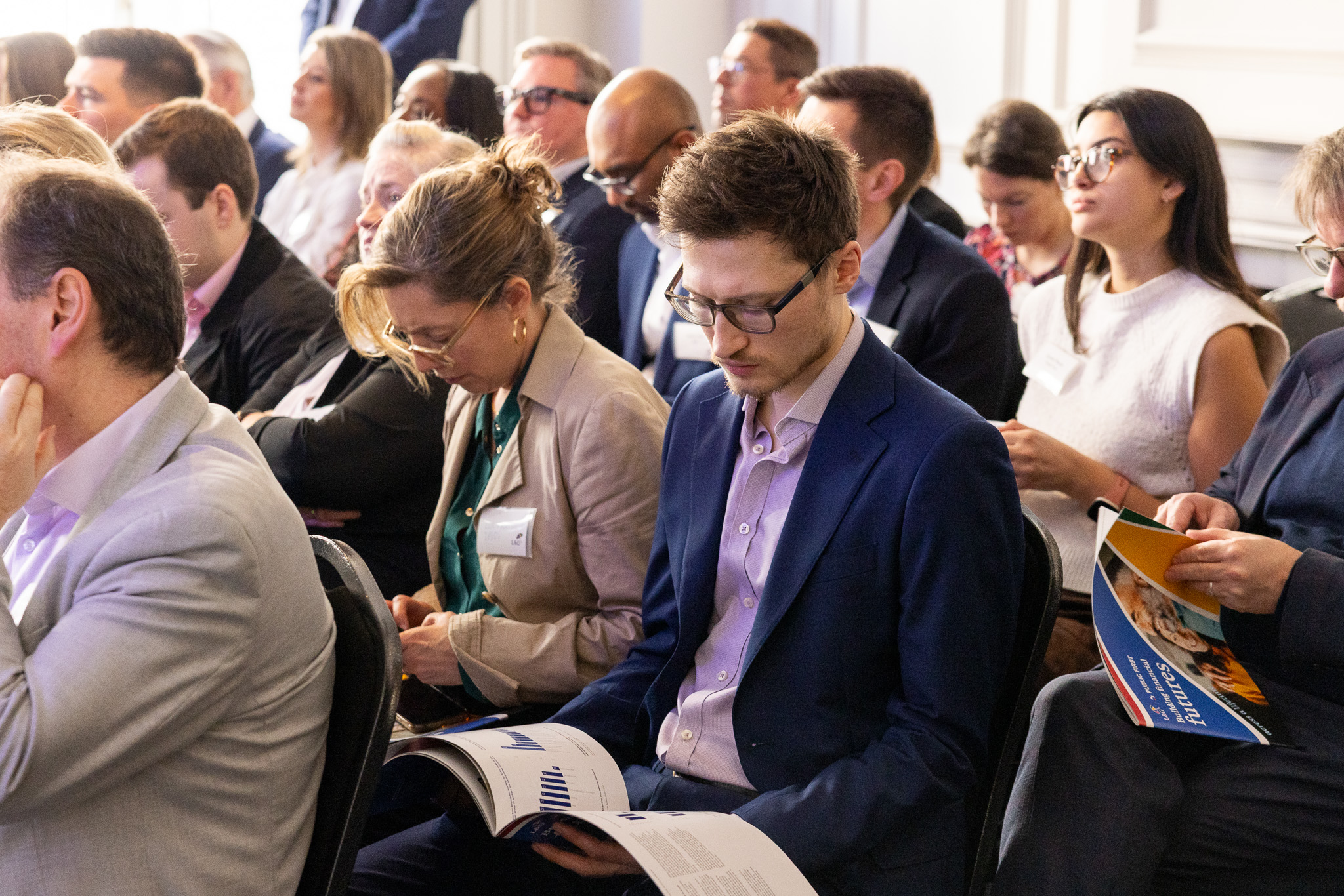 Audience seated in rows in a bright event room holding brochures and listening to a presentation.