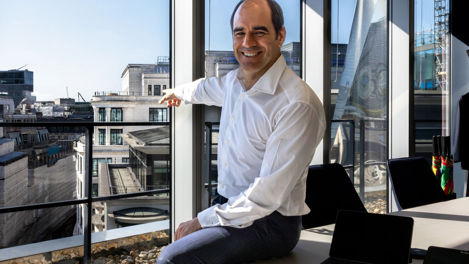 António Simões sitting on a desk in L&G's office at 1 Coleman Street, with a large office window behind him, pointing towards 10 Coleman Street. António Simões sitting on a desk in L&G's office at 1 Coleman Street, with a large office window behind him, pointing towards 10 Coleman Street.