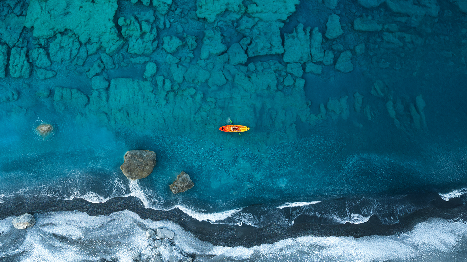 Aerial drone view of a lone kayak gliding over the turquoise waters of Agios Pavlos beach, Crete, Greece, surrounded by coastline.