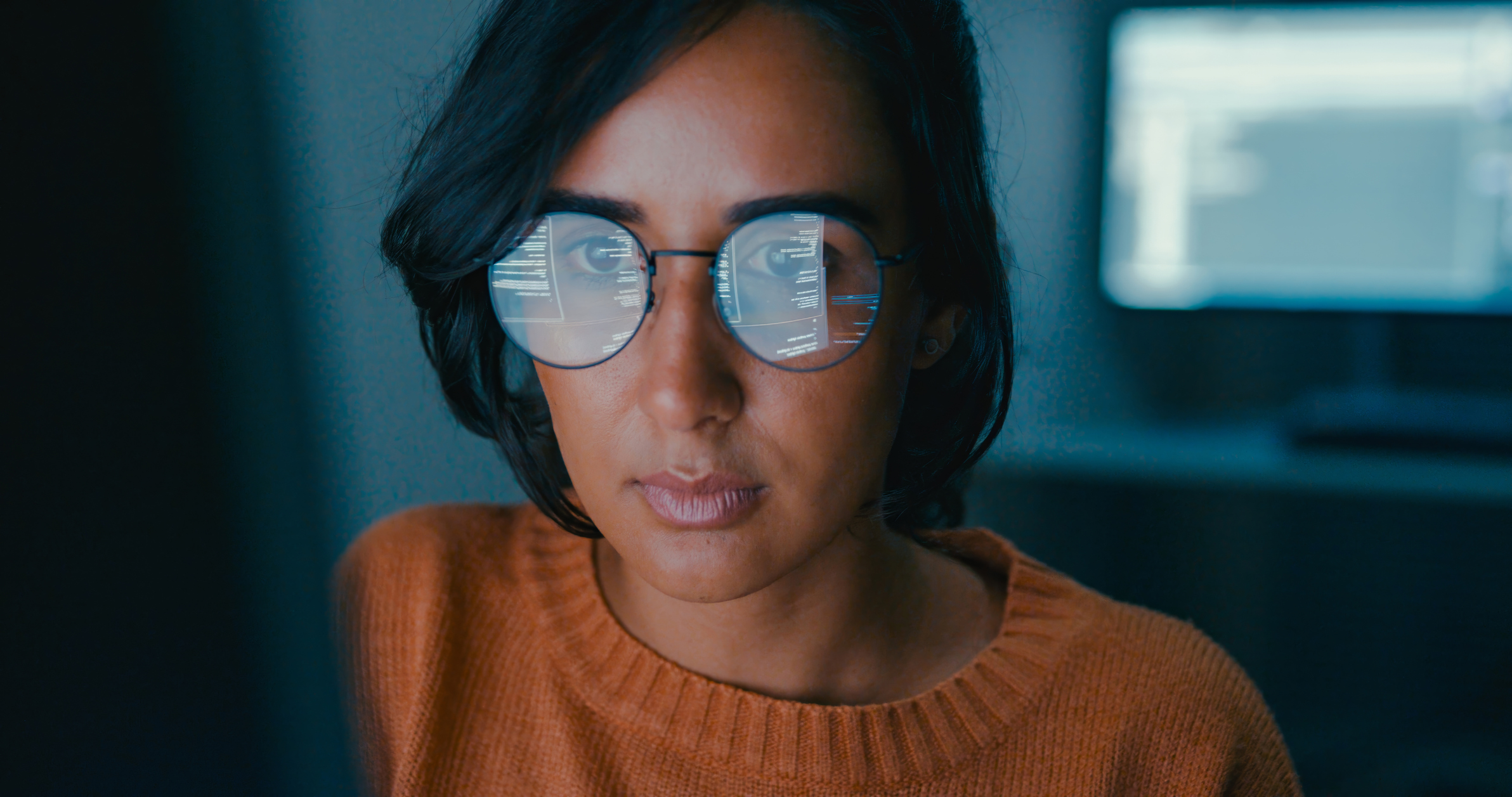 Person sitting indoors working at a computer, with the reflection of the screen visible in their glasses.