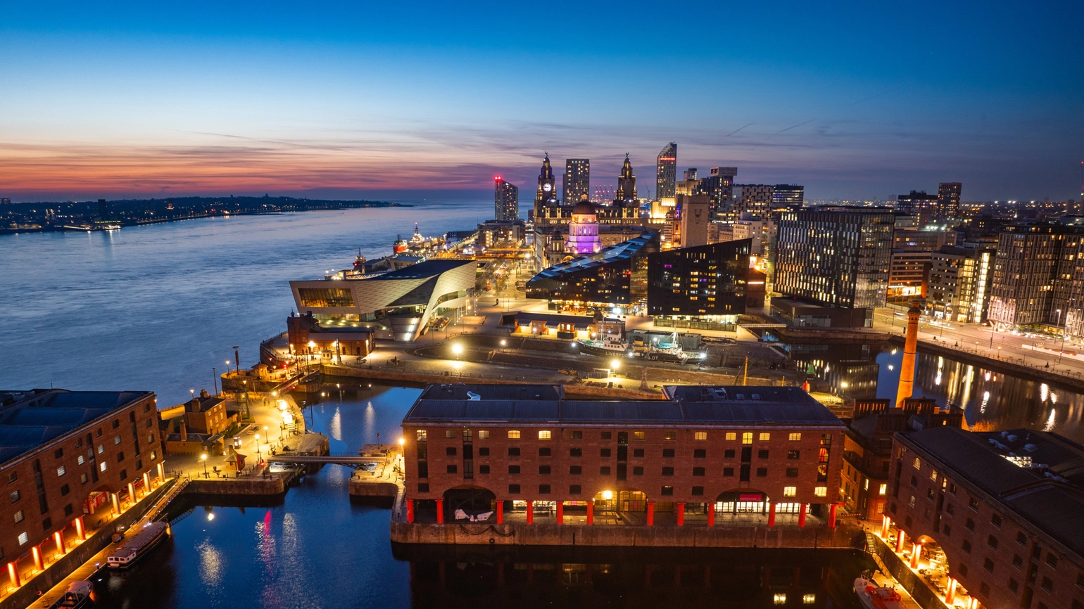 Aerial view of UK city waterfront at dusk, showing illuminated historic dock buildings, modern architecture, and the River Aerial view of UK city waterfront at dusk, showing illuminated historic dock buildings, modern architecture, and the River