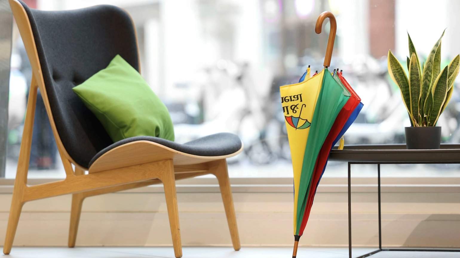 A wooden chair with dark upholstery and a green cushion in front of a large window. Next to the chair is a small black table with a potted plant. On the floor, leaning against the table, is a colourful umbrella with yellow, green, red, and blue panels. The Legal & General logo is visible on the yellow panel. A wooden chair with dark upholstery and a green cushion in front of a large window. Next to the chair is a small black table with a potted plant. On the floor, leaning against the table, is a colourful umbrella with yellow, green, red, and blue panels. The Legal & General logo is visible on the yellow panel.