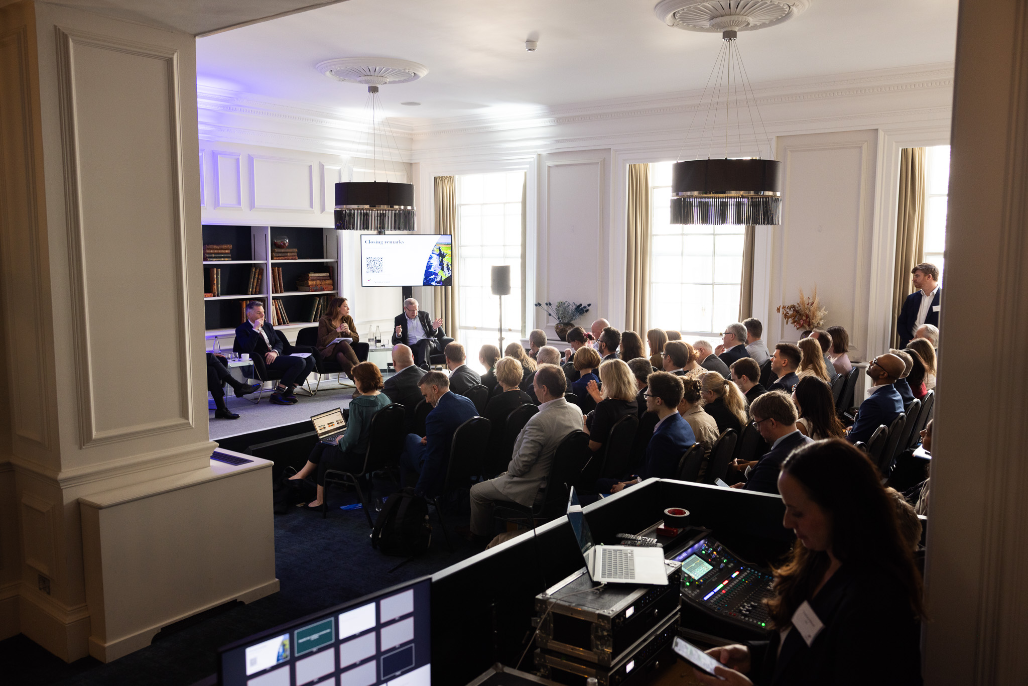 Wide view of a conference room showing the audience, panel speakers, and technical equipment at the back.