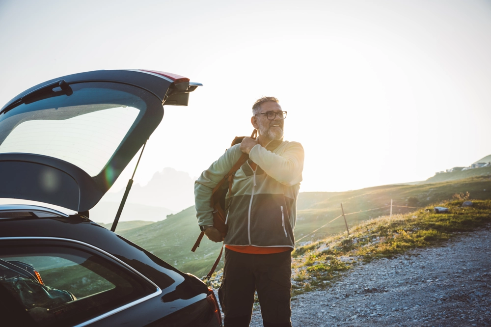 Person standing beside an open car trunk on a gravel path, adjusting a backpack strap. The scene features grassy slopes and distant mountain peaks, illuminated by a warm setting sunlight. Person standing beside an open car trunk on a gravel path, adjusting a backpack strap. The scene features grassy slopes and distant mountain peaks, illuminated by a warm setting sunlight.
