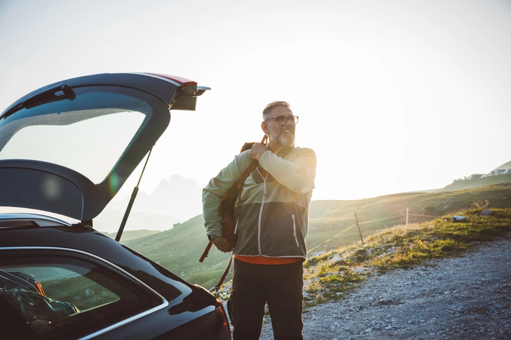 Person standing beside an open car trunk on a gravel path, adjusting a backpack strap. The scene features grassy slopes and distant mountain peaks, illuminated by a warm setting sunlight. Person standing beside an open car trunk on a gravel path, adjusting a backpack strap. The scene features grassy slopes and distant mountain peaks, illuminated by a warm setting sunlight.