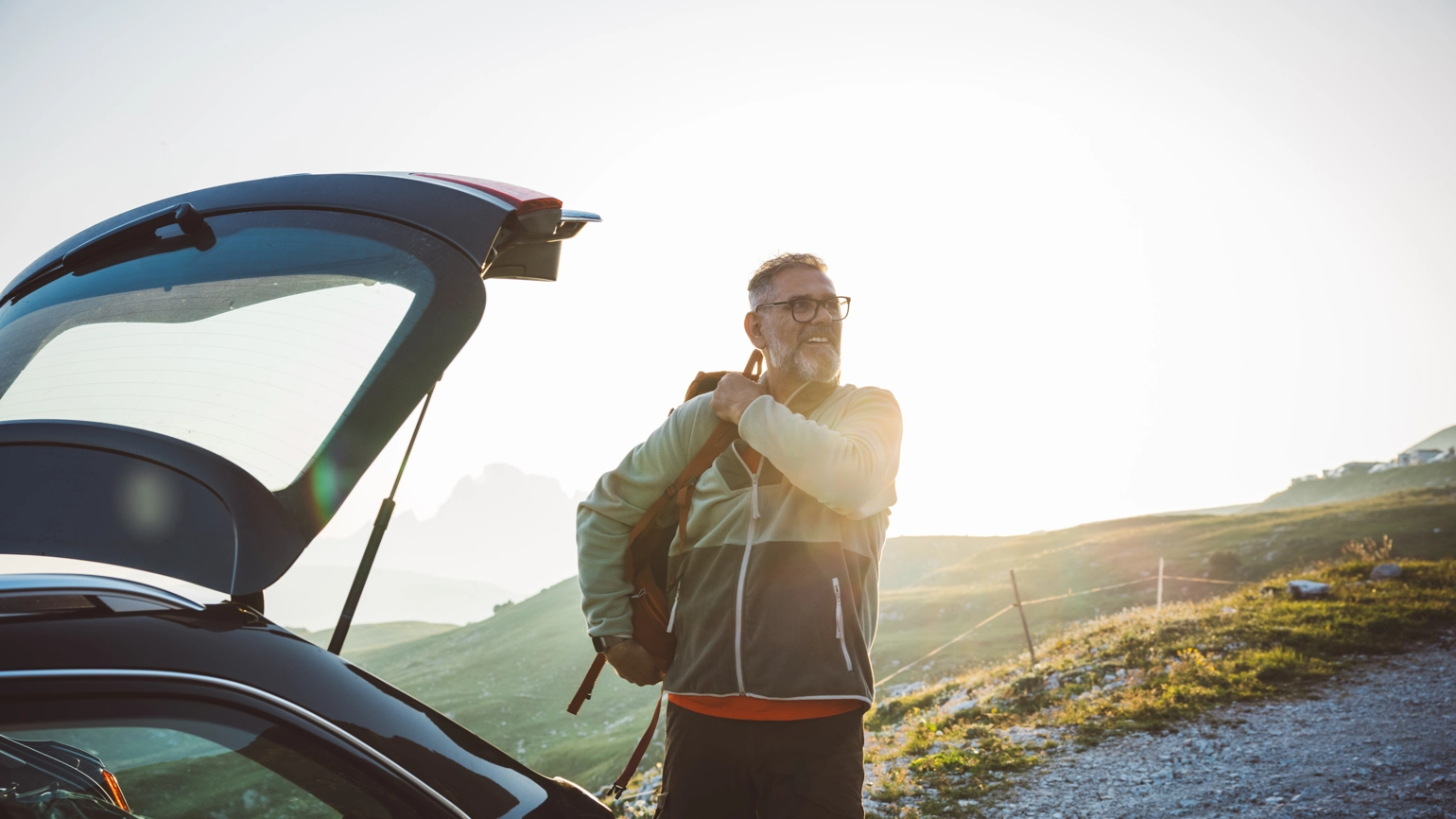 Person standing beside an open car trunk on a gravel path, adjusting a backpack strap. The scene features grassy slopes and distant mountain peaks, illuminated by a warm setting sunlight. Person standing beside an open car trunk on a gravel path, adjusting a backpack strap. The scene features grassy slopes and distant mountain peaks, illuminated by a warm setting sunlight.