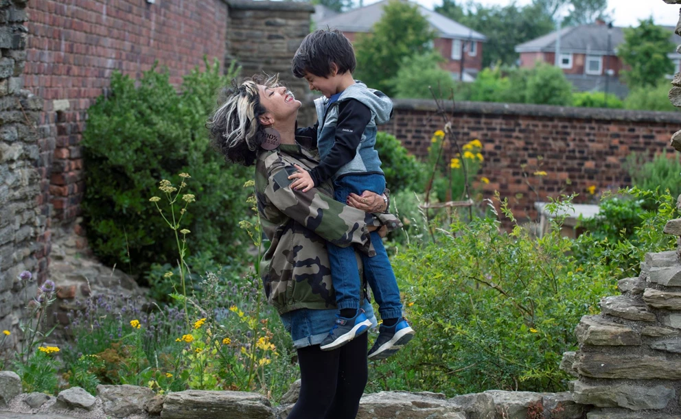 A beaming women carries a child, the pair are in a garden that has wildflowers and rockery 