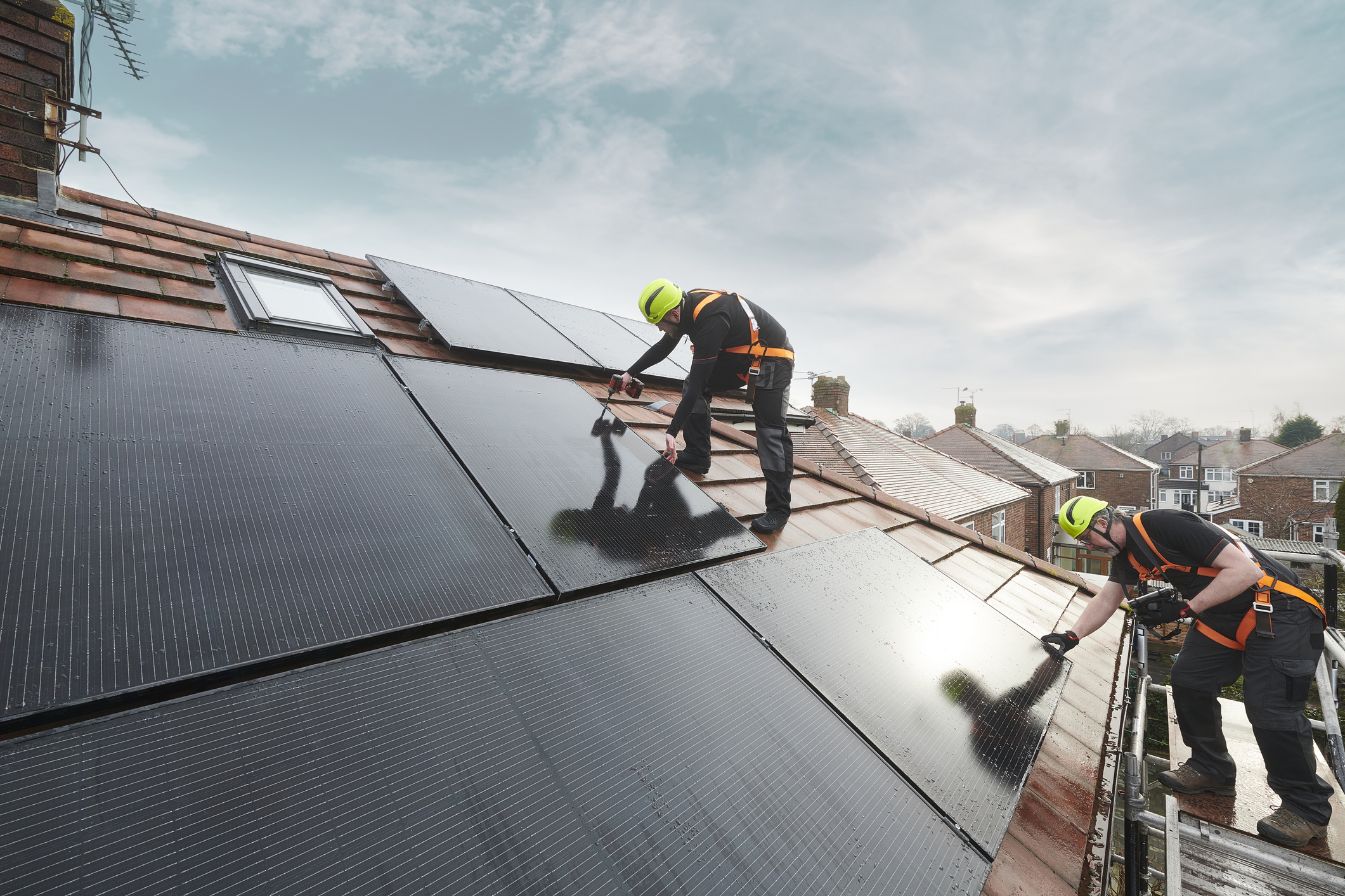 Two workers installing solar panels on a sloped roof, wearing safety harnesses and helmets