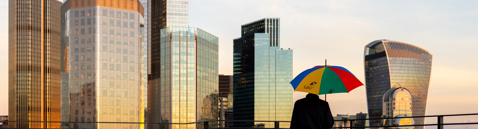 A person viewed from behind stands on a balcony holding a brightly coloured L&G umbrella with red, yellow, green, and blue panels, overlooking a London city skyline of tall modern glass buildings during sunset. A person viewed from behind stands on a balcony holding a brightly coloured L&G umbrella with red, yellow, green, and blue panels, overlooking a London city skyline of tall modern glass buildings during sunset.