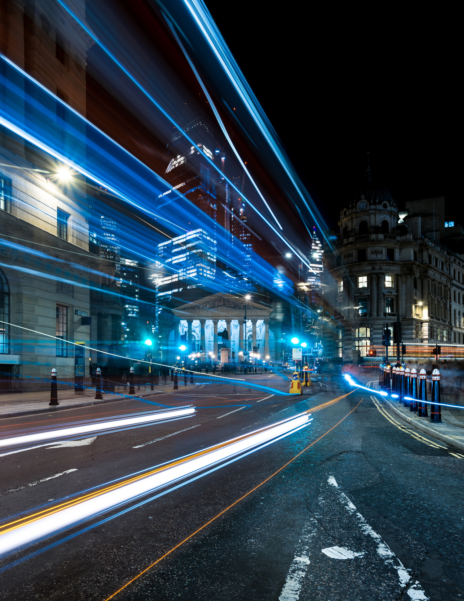 Night view of the Bank Junction in London, UK, with illuminated buildings and skyscrapers in the background. Bright streetlights create starburst effects, and light trails from passing vehicles flow across the foreground.