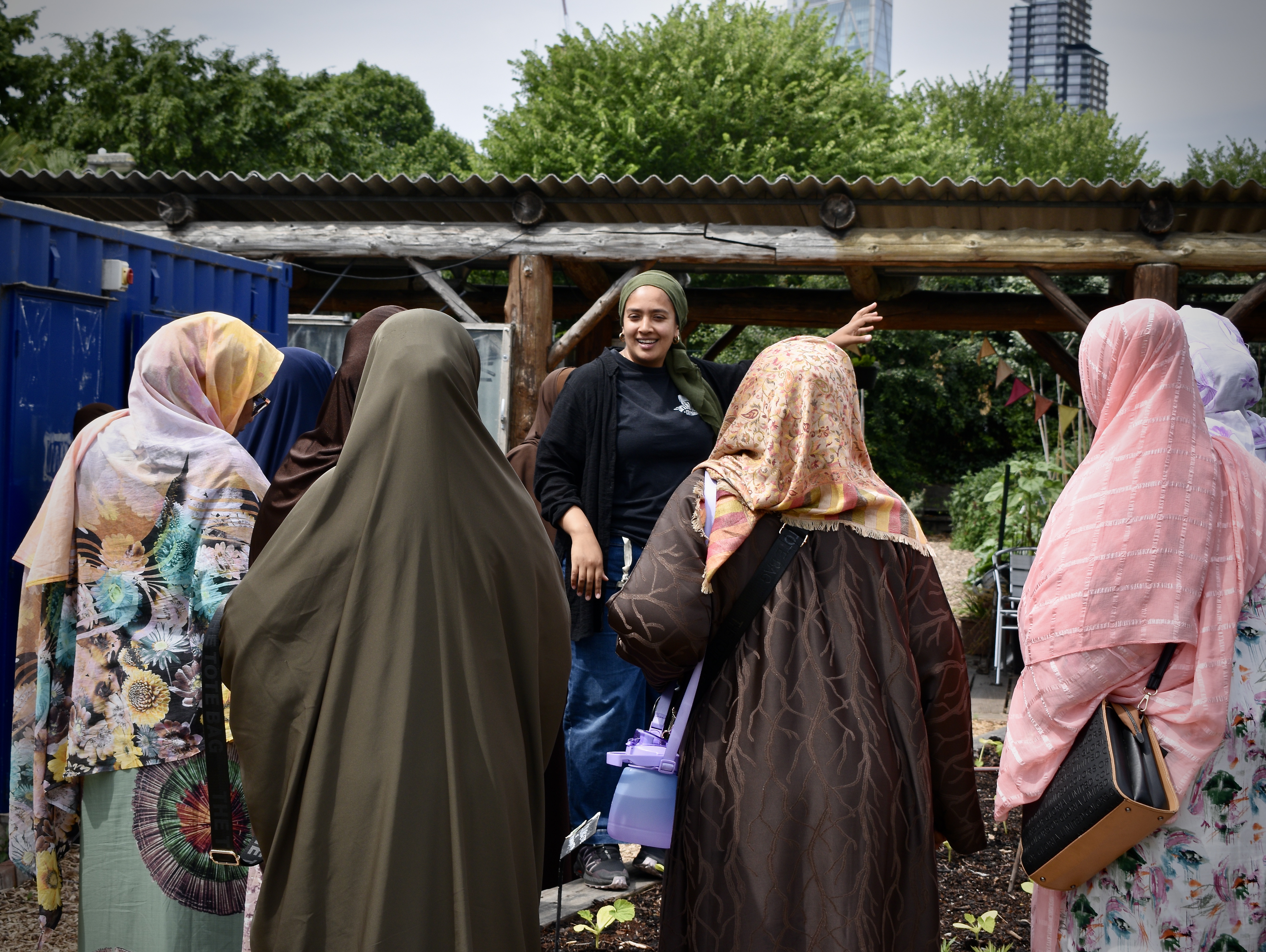 A group of women in hijabs listen to a speaker outdoors, with trees and a wooden structure in the background.