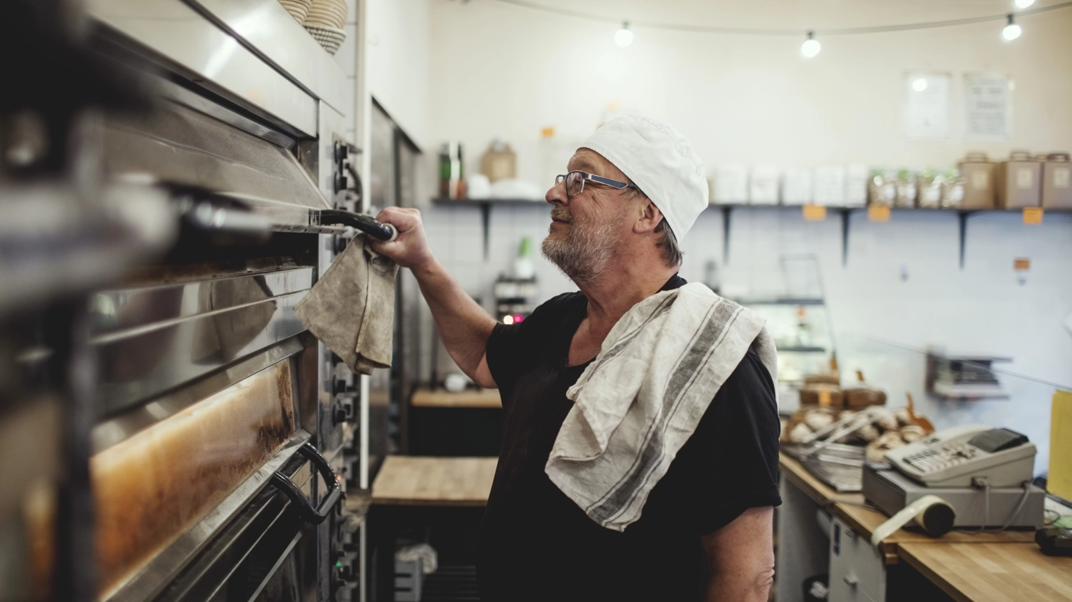 A senior baker stands by a oven at a small bakery. A senior baker stands by a oven at a small bakery.