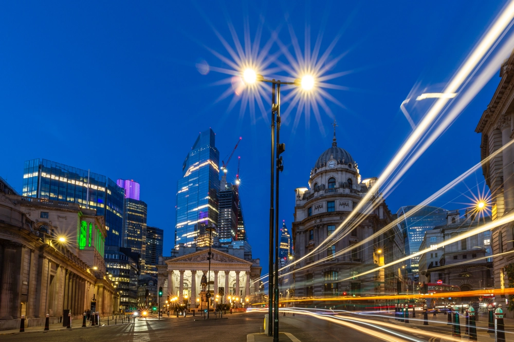 Night view of the Bank Junction in London, UK, with illuminated buildings and skyscrapers in the background. Bright streetlights create starburst effects, and light trails from passing vehicles flow across the foreground. Night view of the Bank Junction in London, UK, with illuminated buildings and skyscrapers in the background. Bright streetlights create starburst effects, and light trails from passing vehicles flow across the foreground.