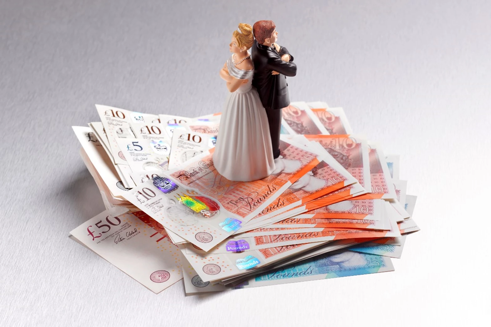A wedding cake topper of a bride and groom, with backs turned and arms crossed, sits on top of a pile of bank notes