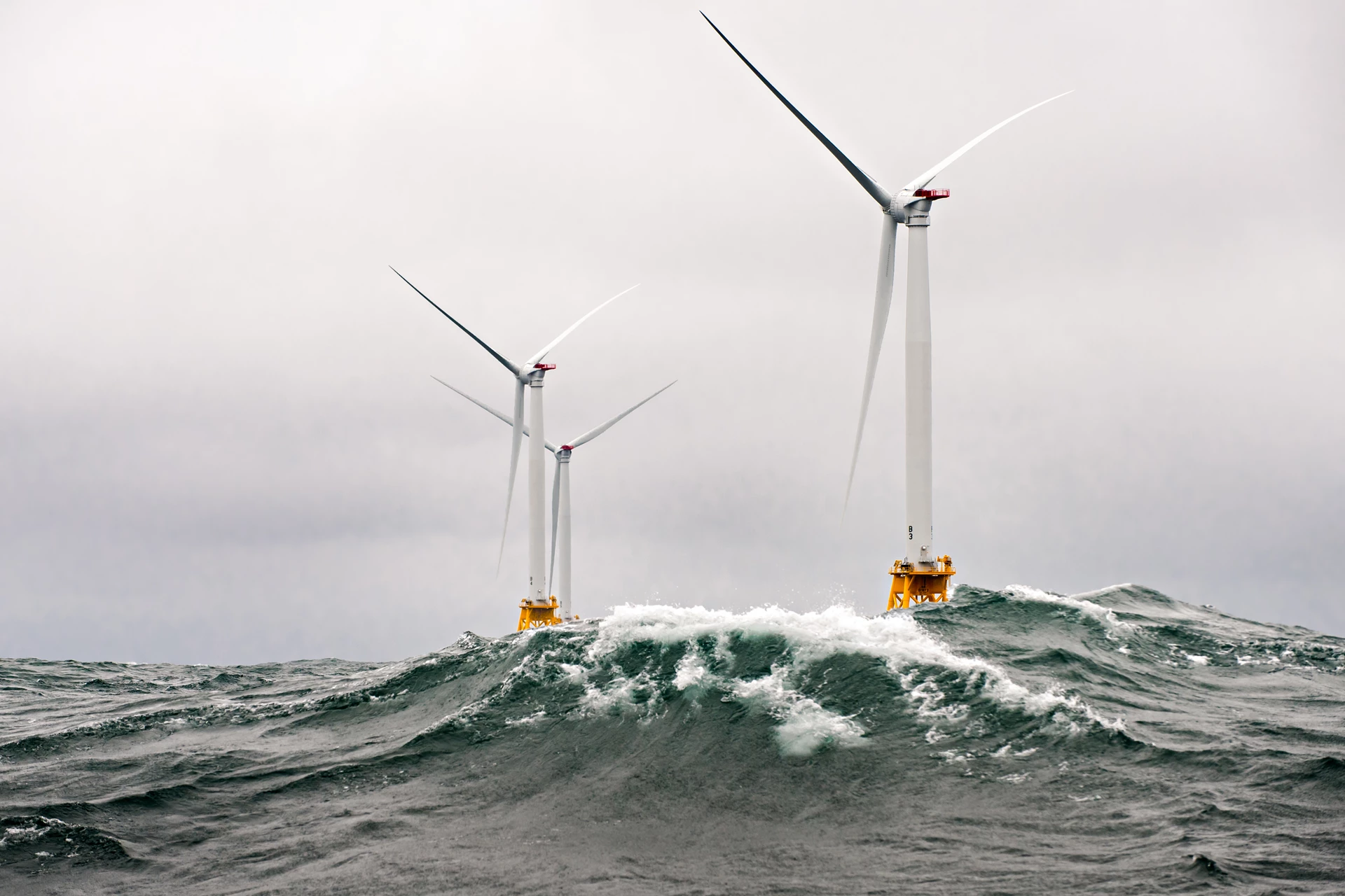 Wind turbines spinning in an off-shore windfarm, with grey skies and waves crashing 