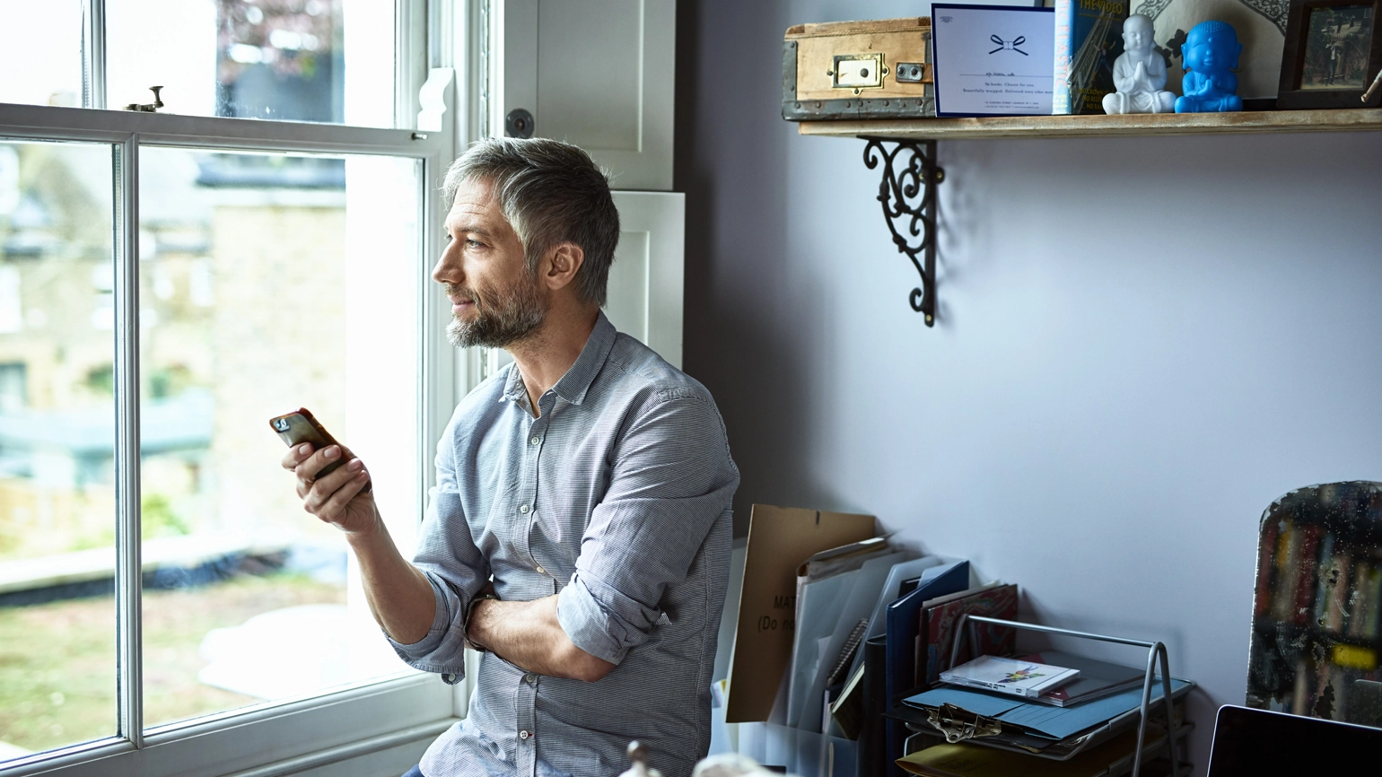 A man is seated by a window, gazing outside while holding a smartphone in his hand. The room is softly illuminated by natural light streaming through the window. A man is seated by a window, gazing outside while holding a smartphone in his hand. The room is softly illuminated by natural light streaming through the window.