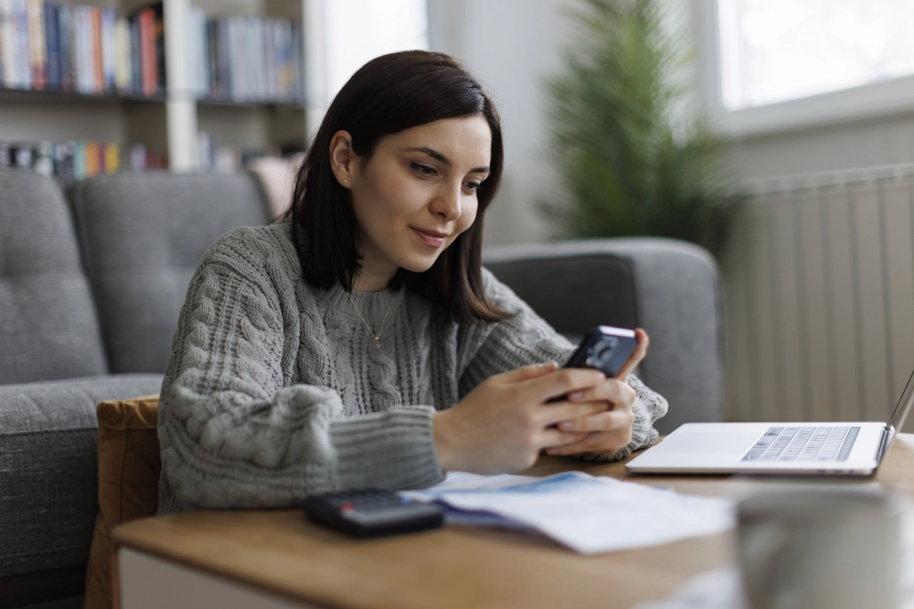 A person leaning on a wooden coffee table in a living room, wearing a grey knitted jumper and holding a smartphone. On the coffee table are scattered papers, a calculator, and an open laptop. In the background, there is a grey sofa, a tall green plant, and shelves filled with books. Daylight comes through a window on the right side. A person leaning on a wooden coffee table in a living room, wearing a grey knitted jumper and holding a smartphone. On the coffee table are scattered papers, a calculator, and an open laptop. In the background, there is a grey sofa, a tall green plant, and shelves filled with books. Daylight comes through a window on the right side.