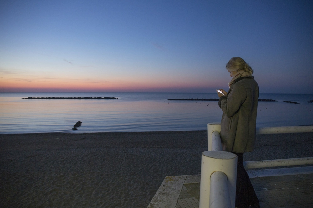 A person wearing a winter coat standing on a seaside promenade at dusk, illuminated by the light of their phone while facing a calm ocean and a sky that transitions from deep blue to soft pink near the horizon. A person wearing a winter coat standing on a seaside promenade at dusk, illuminated by the light of their phone while facing a calm ocean and a sky that transitions from deep blue to soft pink near the horizon.
